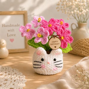 May include: A handmade crochet cat-shaped flower pot filled with pink flowers and green leaves. The pot has black whiskers and pink ears. A wooden tag with "Happy Birthday" hangs from the flowers. The background includes a framed picture that says "Handmade with Love".