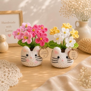Op de afbeelding: Twee handgemaakte gehaakte bloempotten in de vorm van katten, elk met kleurrijke bloemen. Eén pot heeft roze bloemen, de andere witte en gele bloemen. Elke pot heeft een houten label met "Happy Birthday" en "I Best Mom Ever".