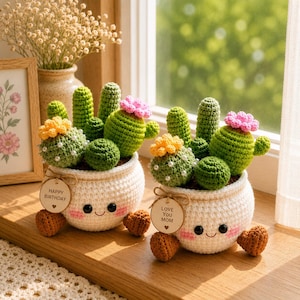 Peut inclure: Deux cactus crochetés dans des pots blancs avec des visages souriants, des cactus verts et des fleurs roses et jaunes. Un pot porte une étiquette avec "Happy Birthday", l'autre "Love You Mom". Accents en forme de cœur marron.