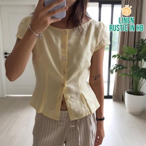 Puede incluir: Blusa de lino amarillo pálido con mangas cortas y botones delanteros. La blusa tiene cintura entallada y pliegues en la parte inferior. El texto "LINEN RUSTIC IN HB" es visible en la esquina superior derecha.