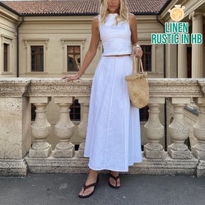 Puede incluir: Conjunto de dos piezas de lino blanco, compuesto por un top sin mangas y una falda larga. El conjunto se combina con sandalias marrones y un bolso tejido. Incluye el texto "LINEN RUSTIC IN HB".