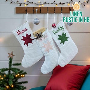 May include: Three white linen Christmas stockings hang from a wooden rack. Each stocking has a star design and a name embroidered on it: "Mama," "Leah," and "Daddy." The stockings are decorated with red, pink, and green stars.