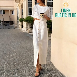 May include: A white linen two-piece set, comprising a cropped top with short sleeves and a high-waisted, button-through maxi skirt with a front split. The outfit is worn with sandals. The image includes the text "LINEN RUSTIC IN HB".