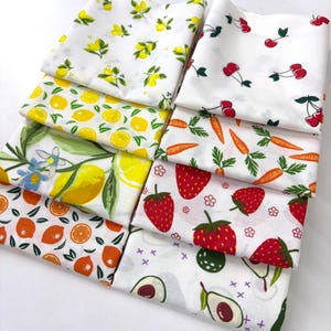 May include: A collection of white fabric squares with various fruit and vegetable patterns. Designs include lemons, cherries, carrots, strawberries, oranges, and avocados. The fabrics are folded and stacked, showcasing the colorful prints.