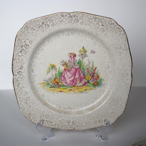 May include: A white square plate with a gold rim and a floral design featuring a woman in a pink dress and a birdhouse. The plate has a white background with a gold speckled pattern.
