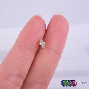 May include: Gold body jewelry with two clear rectangular stones. The jewelry is on a finger.