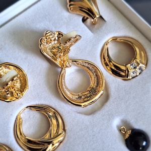 May include: A set of gold-tone earrings with a variety of styles, including a pair of hoop earrings, a pair of clip-on earrings, and a pair of drop earrings. The earrings are all made of metal and have a shiny finish.