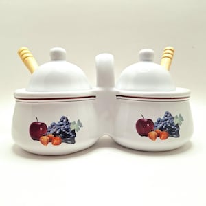 May include: White ceramic double condiment server with lids and wooden spoons. Each side features a fruit design with a red apple, purple grapes, and orange berries. The server has a handle for easy carrying.