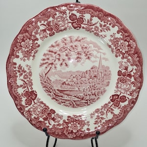 May include: A round, antique-style plate with a scalloped edge. The plate features a red and white color scheme. The center of the plate depicts a rural landscape scene, while the border is adorned with floral patterns. The plate is supported by a black metal stand.