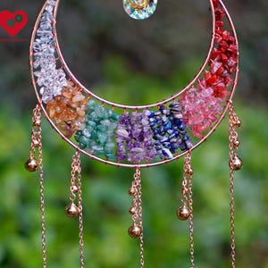 Op de afbeelding: Een halvemaanvormige windgong met een koperkleurig frame, gevuld met kleurrijke edelstenen. De edelstenen zijn gerangschikt in een regenboogpatroon. Goudkleurige kettingen met kleine belletjes hangen aan de onderkant van de maan. Bovenop zit een klein, helder kristal.