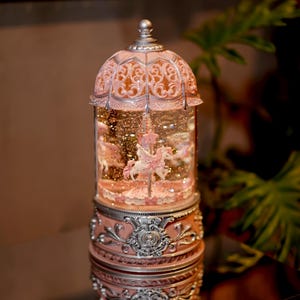 May include: A pink and silver carousel snow globe with a white unicorn and other figures inside. The globe is on a black surface.