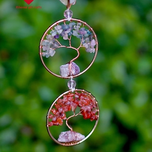 May include: A two-tiered copper-coloured wire tree of life suncatcher. The top circle features purple and green stones, while the bottom circle has red stones. The suncatcher hangs from a clear bead against a blurred green background.