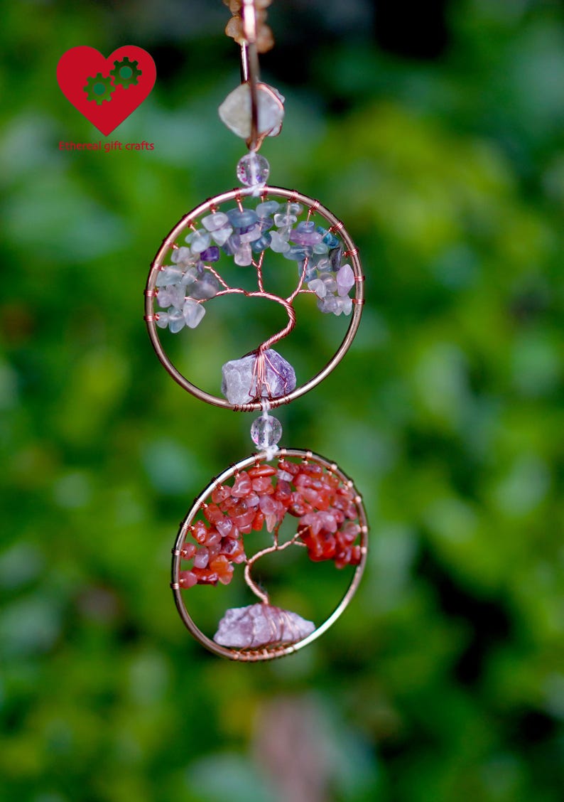 May include: A two-tiered copper-coloured wire tree of life suncatcher. The top circle features purple and green stones, while the bottom circle has red stones. The suncatcher hangs from a clear bead against a blurred green background.