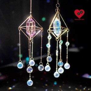 May include: Two hanging crystal suncatchers. Each features a geometric gold frame holding a central crystal, one pink and one clear. Strands of iridescent crystal beads dangle below, creating a shimmering effect. The background is dark with bokeh lights.