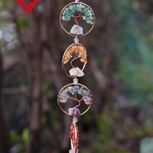 May include: A handcrafted hanging mobile. Three gold-toned circular frames feature wire tree designs and colourful gemstone accents. The top frame has green stones, the middle has orange, and the bottom has purple and red stones. Clear beads suspend the mobile.