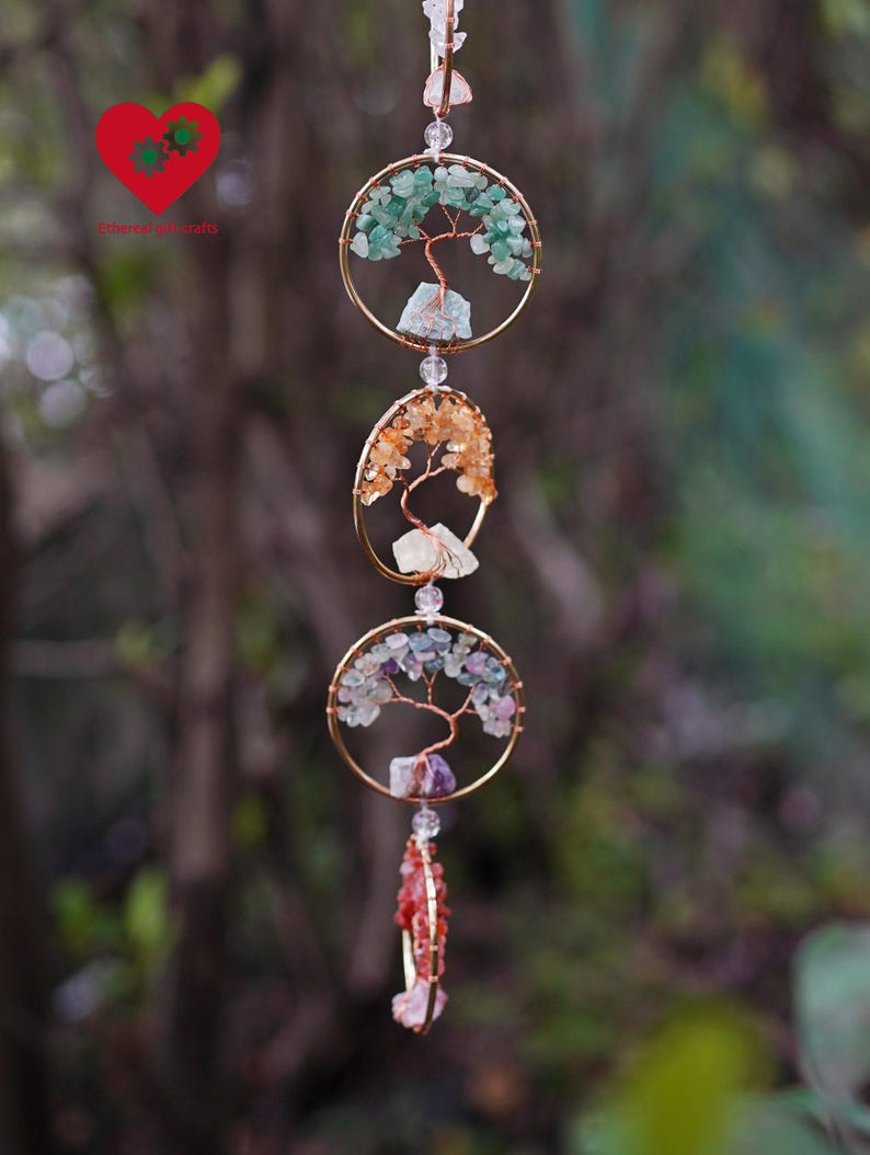 May include: A handcrafted hanging mobile. Three gold-toned circular frames feature wire tree designs and colourful gemstone accents. The top frame has green stones, the middle has orange, and the bottom has purple and red stones. Clear beads suspend the mobile.