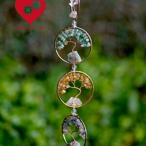 May include: A decorative hanging ornament with three gold-coloured circular frames, each with a wire tree design and gemstone accents. The top frame has green stones, the middle has orange, and the bottom has purple.