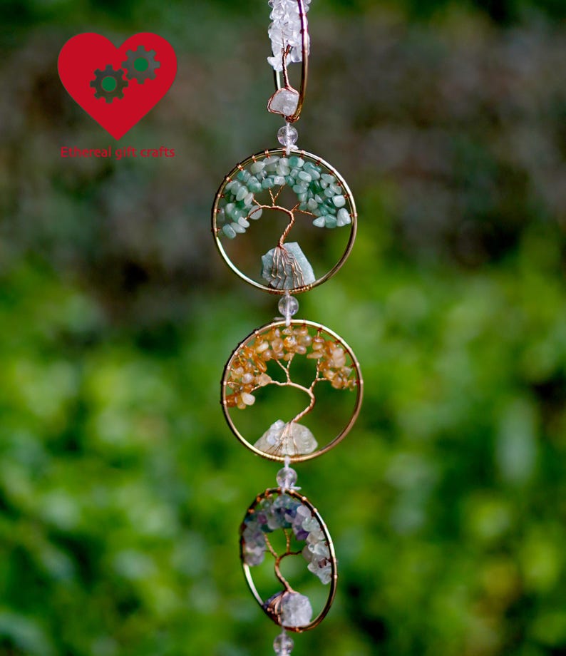 May include: A decorative hanging ornament with three gold-coloured circular frames, each with a wire tree design and gemstone accents. The top frame has green stones, the middle has orange, and the bottom has purple.