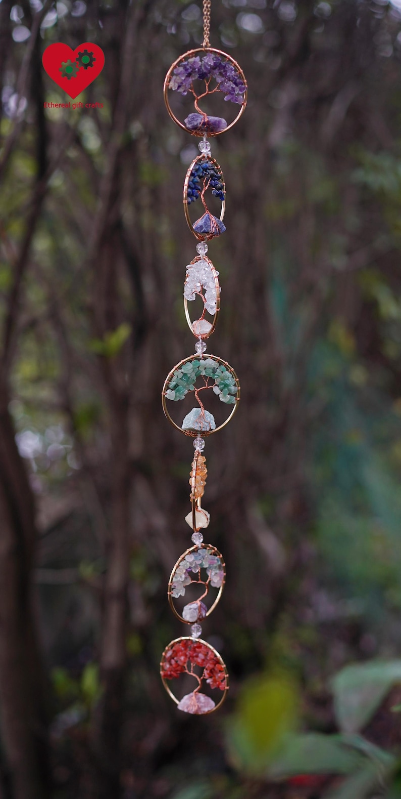 May include: A hanging ornament with gold-coloured circular and oval frames. Each frame features a wire tree design decorated with colourful gemstones. The gemstones include purple, blue, green, and red hues. The ornament hangs from a delicate chain.