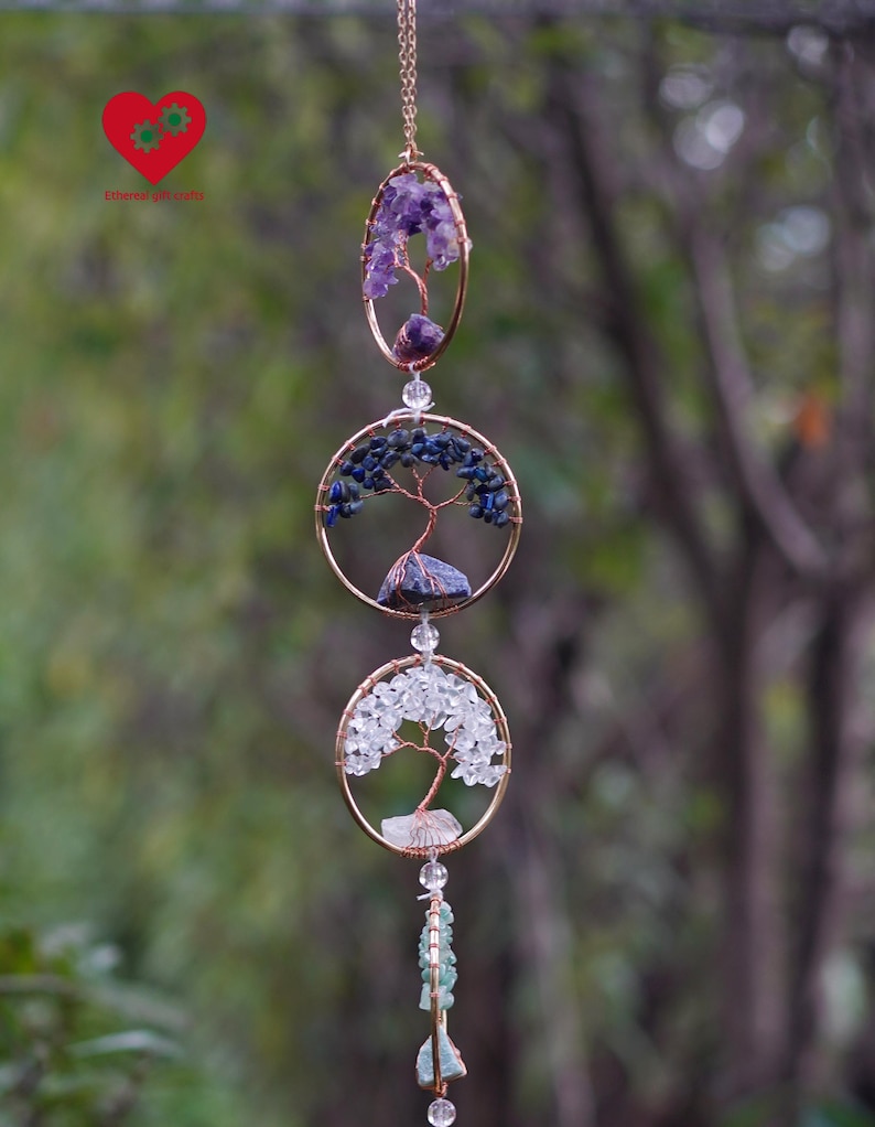 May include: A handcrafted wind chime with three circular and oval frames, each with a wire tree design and filled with amethyst, blue, and clear crystals. The chime is suspended by a gold chain.