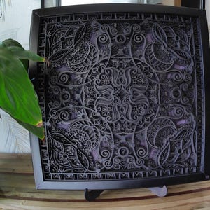 Pode incluir: Uma obra de arte quadrada e em camadas com uma moldura preta. O design intrincado apresenta padrões florais e geométricos em tons de cinza e roxo. A obra de arte é exibida em um suporte, mostrando sua profundidade e detalhes.