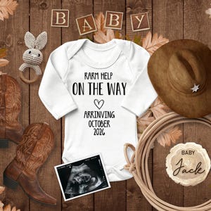 Könnte beinhalten: Ein weißer Strampler mit dem Text "Rarm Help On The Way" und einem Herz mit dem Text "Arriving October 2022". Ein brauner Cowboyhut, ein Paar braune Cowboystiefel, ein Holzspielzeugkaninchen, ein Seil und ein Holzschild mit dem Text "Baby Jack" sind um den Strampler herum angeordnet.