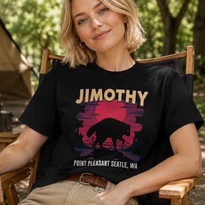 May include: A black short-sleeve t-shirt featuring a graphic print of a dark creature silhouette standing on a rock before a large red sun, a bridge, and trees. The text JIMOTHY is printed above, and POINT PLEASANT SEATLE, WA is printed below.