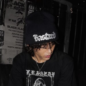 May include: A black knit beanie featuring the word Bastard embroidered in white gothic lettering on the front cuff. The beanie is worn with a black t-shirt that displays a white graphic print of a face and the text Tragedy and Yesterday is Dead.