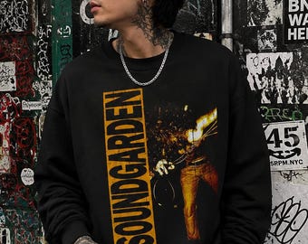 May include: A black long-sleeve sweatshirt featuring a vertical yellow rectangular graphic with the text SOUNDGARDEN in black letters, alongside an illustration of a person holding a flaming object, worn with dark denim jeans and a silver chain.