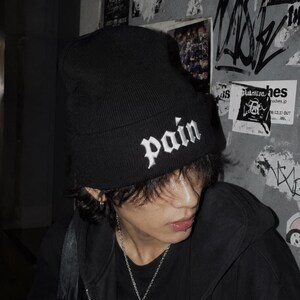 以下が含まれることがあります： 白いスクリプトで「pain」という言葉が入った黒いビーニー。黒いジップアップパーカーと、「walk」という言葉が見える黒いグラフィックTシャツを着ています。首にはシルバーのネックレスがかかっています。