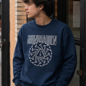 May include: A navy blue crewneck sweatshirt featuring the white text SOUNDGARDEN above a circular graphic design. The graphic consists of a stylized sunburst pattern surrounding a central triangle containing a small spark plug illustration.