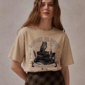 May include: A beige short-sleeve t-shirt featuring a black graphic print of an owl perched on a stack of books, an hourglass, a quill, and an inkwell. The text reads MIDNIGHT OIL SOCIETY, BURNING BRIGHT SINCE 1892, and Sleep is for those who do not.