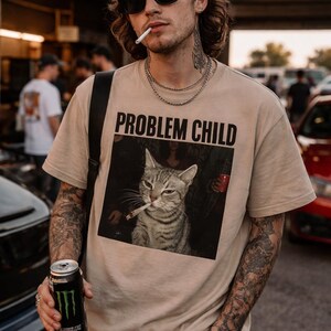 Può includere: Una t-shirt a maniche corte color beige con una stampa grafica di un gatto tigrato con una sigaretta in bocca. Sopra il gatto, il testo "PROBLEM CHILD" è scritto in grassetto nero in maiuscolo. Presenta un girocollo e una vestibilità comoda.