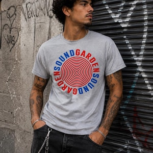 May include: A heather gray short-sleeve t-shirt featuring a circular graphic on the chest. The design consists of a red spiral at the center, surrounded by the text SOUNDGARDEN in alternating blue and red capital letters in a circular layout.