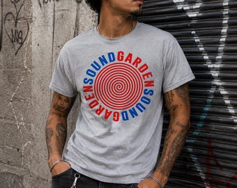 May include: A heather gray short-sleeve t-shirt featuring a circular graphic on the chest. The design consists of a red spiral at the center, surrounded by the text SOUNDGARDEN in alternating blue and red capital letters in a circular layout.