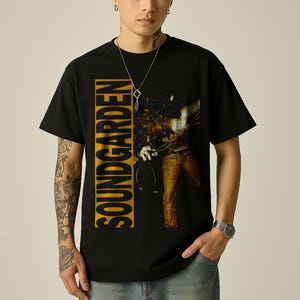 May include: Black t-shirt featuring the band name "SOUNDGARDEN" in yellow vertical text. The shirt has a graphic of a person playing a guitar. The person is wearing jeans and is holding a microphone.