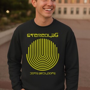 Peut inclure: Sweat-shirt noir avec le texte "Stereolab" au-dessus d'un motif semi-circulaire jaune de lignes verticales et les mots "Dots and Loops" en dessous. Porté par une personne souriante devant un arrière-plan flou.