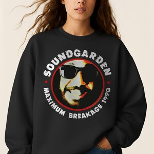 May include: Black sweatshirt featuring a circular graphic with the text "SOUNDGARDEN" and "MAXIMUM BREAKAGE 1990." The graphic includes a portrait of a person wearing sunglasses. The sweatshirt is worn by a person wearing blue jeans.