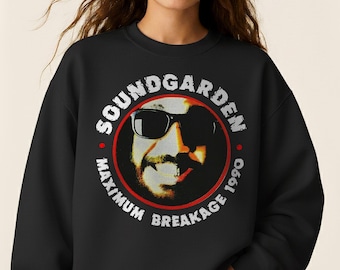 May include: Black sweatshirt featuring a circular graphic with the text "SOUNDGARDEN" and "MAXIMUM BREAKAGE 1990." The graphic includes a portrait of a person wearing sunglasses. The sweatshirt is worn by a person wearing blue jeans.