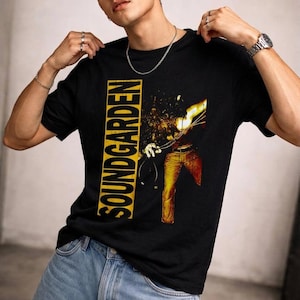 May include: A black short-sleeved t-shirt featuring the word SOUNDGARDEN printed vertically in bold yellow letters inside a rectangular box. To the right, a graphic depicts a figure in yellow pants with a bright, abstract light effect.