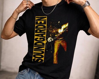 May include: A black short-sleeved t-shirt featuring the word SOUNDGARDEN printed vertically in bold yellow letters inside a rectangular box. To the right, a graphic depicts a figure in yellow pants with a bright, abstract light effect.
