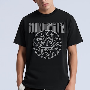 May include: Black t-shirt featuring the band name "SOUNDGARDEN" in white outlined text above a white graphic. The graphic is a stylized triangle with a lighthouse inside, surrounded by a swirling design. The shirt is worn by a person.