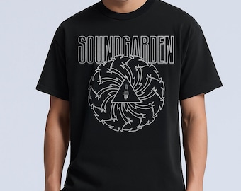 May include: Black t-shirt featuring the band name "SOUNDGARDEN" in white outlined text above a white graphic. The graphic is a stylized triangle with a lighthouse inside, surrounded by a swirling design. The shirt is worn by a person.