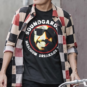 May include: Black t-shirt featuring a graphic of a man wearing sunglasses, encircled by the words "SOUNDGARDEN" and "MAXIMUM BREAKAGE". The shirt is worn under a red, black, and tan checkered button-down shirt.