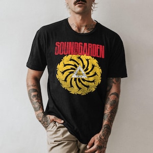 May include: Black t-shirt featuring the band name "SOUNDGARDEN" in red block letters above a yellow and black circular design with a white triangle in the center. The shirt has a vintage, distressed look.