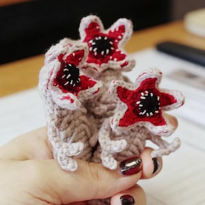 Könnte beinhalten: Handgefertigte Demogorgon-Figuren aus Häkelarbeit. Die Figuren haben rot-schwarze, blütenartige Köpfe mit weißen Akzenten und graue Körper. Die Figuren werden in einer Hand gehalten, was die komplizierten Details des Handwerks zeigt. Inspiriert von einer beliebten Science-Fiction-Serie.