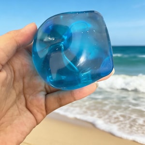May include: A transparent, blue, irregularly shaped glass object is held in a hand against a backdrop of a beach and ocean. The glass has a swirling internal design, and the overall effect is of a cool, aquatic aesthetic.
