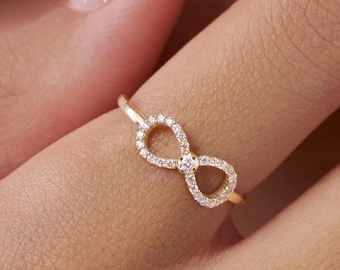 Könnte beinhalten: Ein goldfarbener Ring mit einem Unendlichkeitszeichen-Design. Das Unendlichkeitszeichen ist mit kleinen, klaren Edelsteinen besetzt. Der Ring wird an einem Finger getragen und zeigt sein zartes und elegantes Aussehen. Das Design des Rings ist schlicht, aber auffällig.