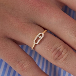 Könnte beinhalten: Ein goldener Ring mit einem kleinen ovalen Design, das mit winzigen Diamanten besetzt ist. Der Ring befindet sich an einem Finger.