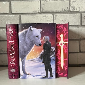 May include: A book cover featuring a white wolf facing a person in a snowy landscape. The book is titled "Dire Bound" by Sable Sorensen. The spine is burgundy with white text. The cover art depicts a fantasy theme.